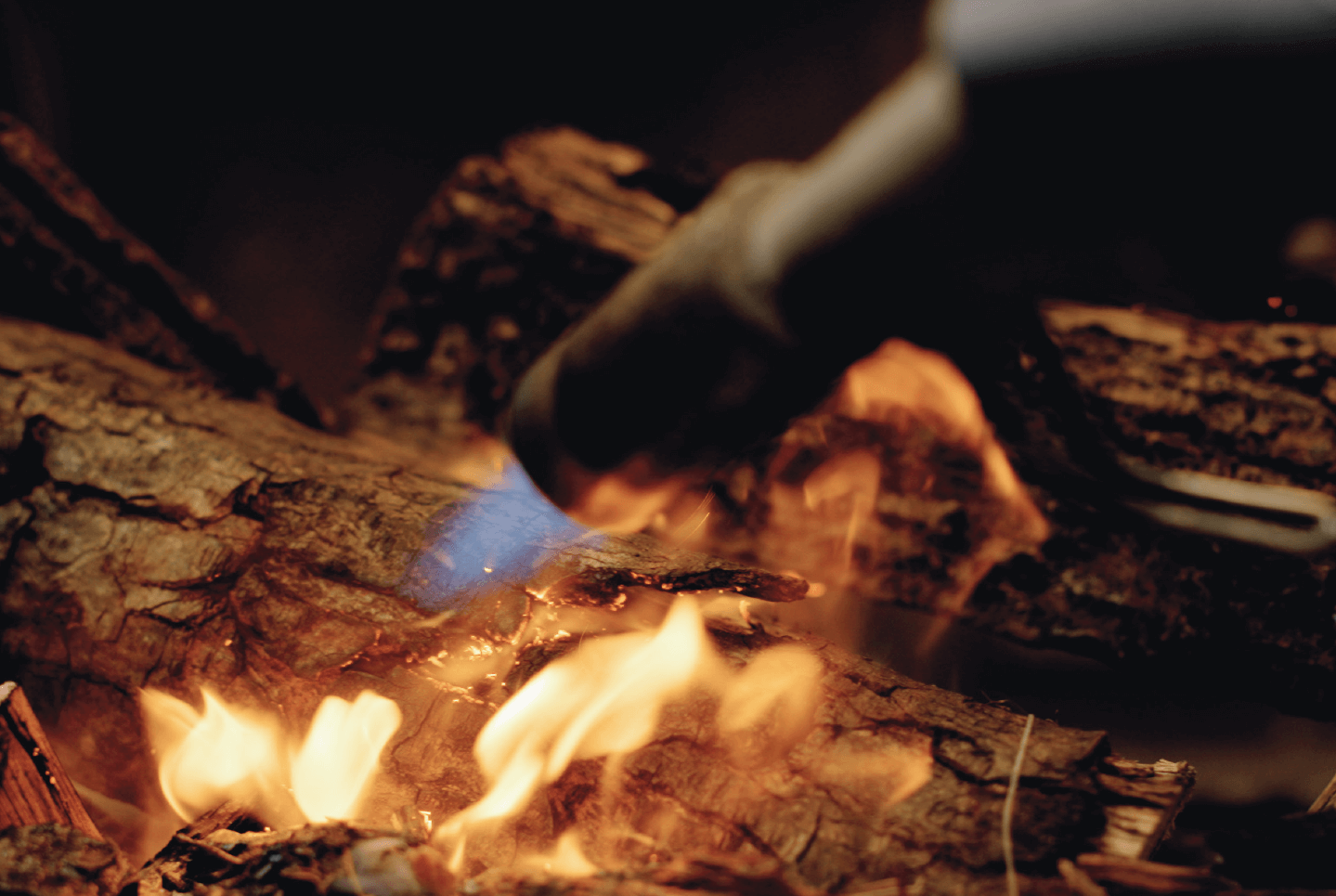 A hand uses a torch to ignite flames on a log in a fire.